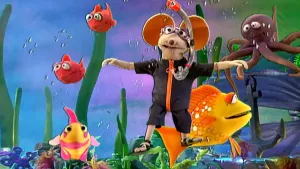Een foto van pop Ieniemienie in een zee met vissenpoppen twee rode, twee gele en een bruinachtige octopus. Ze draagt een donkerpaars zwempak met oranje rits, een paarse duikbril met een snorkel eraan en heeft sandalen met zwarte bandjes om haar voeten. 