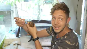 Een foto van Elbert die in de camera kijkt. Hij heeft in zijn linkerhand een kleine, bruine Eekhoorn vast. In zijn rechterhand heeft hij een kleine spuit, waarop hij drukt met zijn vinger. Aan de spuit zit een drinkmondje, waar de Eekhoorn uit drinkt.