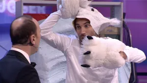 Een foto van een presentator en een man in witte kleding: een wit shirt met lange mouwen, een ijsberenmasker die hij omhoogdoet en om zijn handen een soort sloffen met zwarte klauwen. Achter hem staat een soort neppe lichtblauwe steen of ijsschots.  