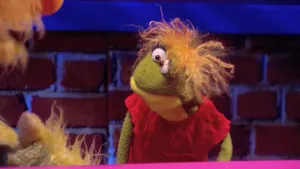 Een foto van een pop die Benthe heet. Benthe heeft een groene huid die geel is onder de kin, naar elkaar toestaande, ronde ogen, oranje haar en een rode jurk. Links is een stukje van het gezicht en de hand van de gele Opper de Pop te zien.