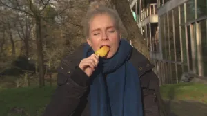 Een foto van Janouk die met haar ogen dicht buiten staat en een ijsje in haar mond steekt. Het is een waterijsje van het type rakket, waarvan de gele en oranje delen te zien zijn. Janouk draagt een donkergrijze jas en een blauwgrijze sjaal.