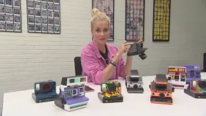 Een foto van Janouk die achter een witte tafel zit en een directklaarcamera vastheeft en ernaar wijst. Op de tafel staan nog acht directklaarcamera's allemaal van verschillende kleuren. Bij het maken van een foto hiermee komt de foto direct uit de camera.