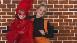 Een foto van een muur met daarvoor Janouk in oranje broek met banden om haar schouders en een zwart shirt. Ernaast staat een vrouw in een rood pak met rode veren en vormen van vlammen erop, een rode muts van veren en een zonnebril met rode glazen op. 