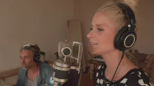 Een foto van Janouk die een hoofdtelefoon opheeft en zingt in een grote microfoon die vóór haar staat. Erachter zit een man met een hoofdtelefoon op, die de muziek opneemt en bewerkt op een niet-zichtbare computer.