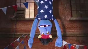 Een foto van een pop die naar beneden duikt. De pop heeft een blauwe huid, zwart haar, een rode band met witte stippen en twee hoorns erop om zijn hoofd en een blauw t-shirt met witte sterren. Erachter hangen op een stenen gebouw rood-wit-blauwe slingers.