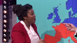 Een foto van weervrouw Wietske Storm die voor een lichtblauwe kaart staat waar de landen linksboven in Europa, onder andere Nederland, donkerblauw zijn en de landen linksonder van Europa zijn rood gekleurd, omdat het daar meestal warmer is.