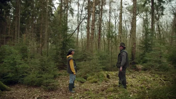 Een foto van een bos met bomen met lange stammen en dennenbomen met groene takken. Vooraan op een gedeelte waar nog geen bomen staan, staan Manon met een groene pet,  mouwloze jas en laarzen en een boswachter die helemaal groen gekleed is, inclusief hoed.