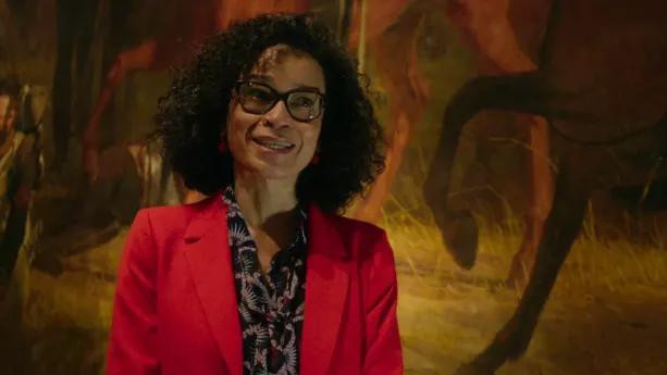 Een foto van een vrouw voor het schilderij "De Slag bij Waterloo", dat voor een klein deel te zien is. Te zien zijn de benen van twee paarden, allebei één been geheven en lopend op een pad. De vrouw draagt een rood jasje en een donkere blouse met print.