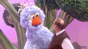 Een foto van pop Pino, met lichtblauwe veren, oranje snavel en grote, ronde ogen. Naast hem staat Buurman Baasje en ze kijken naar elkaar. Buurman Baasje heeft een bruin vest aan en een witte blouse met ruitjes. Erachter staan stronken groene broccoli.