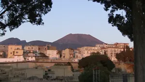 Een foto van witte en lichtbruine huizen met enkele bomen vooraan. Achter de huizen is de top van een paarsachtig gekleurde berg, een vulkaan, te zien. 