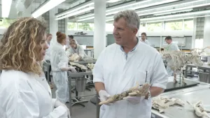 Een foto van Rachel en Arend in een witte jas. Arend heeft een dood biggetje vast die wit van kleur geworden is. Erachter zijn andere mensen ook met skeletten of dode dieren aan het werk op metalen tafels. 