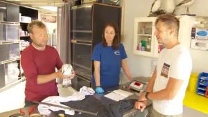 Een foto van twee mensen van de vogelopvang en Elbert. De man heeft een witte meeuw vast, die schoongemaakt is en onder het frituurvet zat. Er staan kasten met doeken en verzorgingsmiddelen erin en op een tafel staat een weegschaal en liggen doeken.