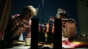 Een foto van een donkere ruimte met een tafel waarop een bak staat met zwarte randen en water erin. Sosha raakt één van de rode strookjes crêpepapier in het water aan, gele strookjes liggen er ook in. Rogier heeft een geel rietje en plastic bekertje vast.