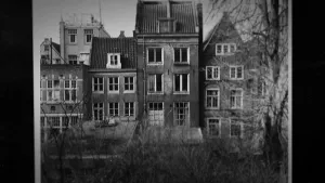 Een zwart-wit foto van een huis tussen twee anderen huizen in.  Het is de Prinsengracht 263 in Amsterdam, het achterhuis waar de Joodse Anne Frank en haar familie ondergedoken zaten voor de nazi's. Het huis heeft zes ramen en een schuin dak.