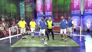 Een foto van een klein voetbalveld met een groen, rechthoekig tapijt  met witte lijnen erop en twee witte doelen in een televisiestudio. Er lopen drie mannen met gele hesjes en drie mannen met blauwe hesjes op en er ligt een bal. Ze spelen wandelvoetbal.