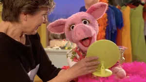 Een foto van Lot die een groene, plastic spiegel vastheeft en die voor de pop van varken Purk houdt. Purk kijkt in de spiegel en heeft haar bek open. Purk draagt een witte jurk met roze rozen en er ligt een boa van roze veren, een soort sjaal, vóór Purk.