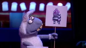 Een foto van pop Stella Schaap met een bord waarop een getekende boef in zwart-wit gestreept pak en muts staat. Ze kijkt ernaar. De pop heeft een witte vacht op rug en oren, een grijze vacht bij kop en poten, ronde ogen en vooruitstekende, gele tanden.