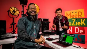 Een foto van Jeangu Macrooy met een koptelefoon om zijn nek achter een bureau met apparaten met draai- en drukknopjes. Naast hem staat een laptop computer waarop in een programma witte, kronkelende lijnen op groene vlakken staan. Buddy staat erachter.