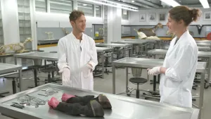 Een foto van Elbert en leraar Luciel die in witte jas en met witte, plastic handschoenen aan bij een stalen tafel staan, een snijtafel. Elbert wijst met zijn handen naar twee zwarte koeienpoten die op de tafel liggen, naast scharen en mesjes.  