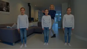 Een foto van drie meisjes die er precies hetzelfde uitzien en naar de camera kijken. Het is een eeneiige drieling. Ze dragen allemaal een witte trui, een blauwe spijkerbroek en witte sokken. Janouk staat achter het middelste meisje in de woonkamer.