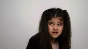 Een foto van een meisje voor een grijze achtergrond. Het meisje kijkt met een verdrietige blik naar de linker bovenkant van de foto. Het meisje heeft lang, donker haar dat aan beide kanten op haar schouders hangt.