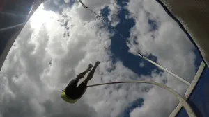Een foto vanaf de grond genomen van iemand die met een lange, gebogen stok een sprong aan het maken is en probeert over een lat te springen. Diegene doet aan polsstokhoogspringen. De blauwe lucht met witte wolken is zichtbaar erboven.