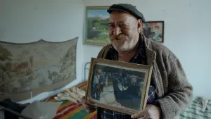 Een foto van de Bulgaarse Radi in een vest en met pet op, die naast een bed staat. Hij houdt een foto vast met zichzelf en een bruine beer erop die hij aan touwen vastheeft die aan banden op de kop van de beer vastzitten. De beer staat op de achterpoten. 