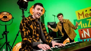 Een foto van Duncan Laurence en Buddy. Duncan staat achter een elektrische piano. Buddy houdt zijn handen naast zich omhoog en kijkt naar de camera. Achter hen staan drie lampen voor op een podium, een microfoon en versterkers om geluid harder te maken.