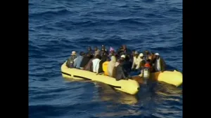 Een foto van een gele motorboot in het water van de middellandse zee in 2004. Er zit een groep van 35 Afrikaanse vluchtelingen op de boot, waaronder Aziz, die de Italiaanse kust willen bereiken. 