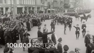 Een zwart-wit foto met mensen in een donkerkleurig legerkostuum. Links staan mensen toe te kijken en staat iemand in een auto die de nazigroet doet, rechts lopen mensen los en in een rij en zitten twee mensen op een paard. Ervoor staat: "100.000 NSB'ers".