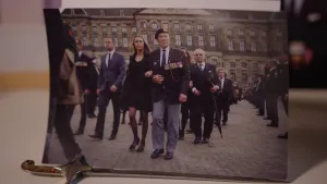 Een foto met een foto erop die op een witte tafel of kastplank staat. Op die foto lopen mensen op de Dam in Amsterdam tijdens dodenherdenking. Rudi Hemnes loopt vooraan in een legerkostuum met pet en veel gekleurde lintjes met medailles op zijn borst.