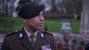 Een foto van soldaat Jackson Rodrigues da Cruz. Hij draagt donkerpaarse kleding: een pak een stropdas op een witte blouse en een pet. Op één schouder hangt een blauw met oranje gevlochten versiering. Op zijn borst hangen drie medailles.