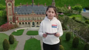 Een foto van het meisje Ileen met een kaartje in haar handen. Ze draagt een trui met de tekst "Nationale Kinder Herdenking 4 Mei" erop. Erachter is het vredespaleis in Den Haag te zien. In het paleis worden internationale ruzies opgelost.