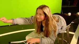 Een foto van een meisje dat gestrekt op een strijkplank ligt met haar rechtervuist naar voren. De strijkplank heeft een hoes met witte en groene driehoeken erop. De strijkplank staat op twee grijze, houten krukken. Achteraan hangt een groen scherm.