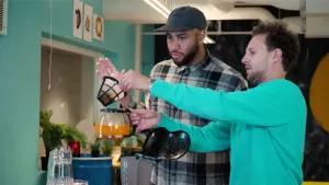 Een foto van Edson en Tim. Tim heeft een glazen kan van een koffiezetapparaat vast, waarin oranje gekleurde soep zit. Hij haalt de filter van het koffiezetapparaat uit de opening bovenop.