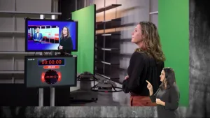 Een foto van gebarentolk Irma voor een rechthoekig, groen vlak. Ze kijkt naar een beeldscherm van een live televisieuitzending met een presentator en zijzelf. Eronder hangt een beeldscherm met 8 uur 's avonds erop. Een andere gebarentolk geeft uitleg.