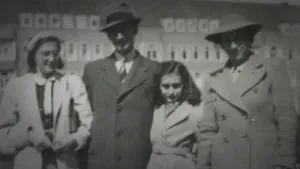 Een zwart-wit foto met de familie van Anne Frank. Links haar zus Margot, daarnaast haar vader Otto, dan Anne zelf en dan haar moeder Edith. Ze dragen een lange, dichtgeknoopte jas en behalve Anne heeft iedereen een hoed op. Achteraan staat een gebouw.