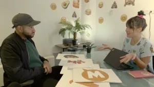 Een foto van Edson en Lilian achter een glazen tafel met daarop vellen papier met niet-bestaande emoji's: een W in een gele tekstballon, het lachende gezicht van Edson, een clownsvis en stroopwafels zijn te zien. Op de muur zitten stickers van emoji's.