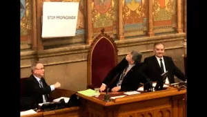 Een foto van drie mannen achter een hoge tafel. De middelste man zit achter een hoge, houten stoel, bekleed met rode stof op de rug. Hij kijkt omhoog richting de muur met schilderingen erop achter hem, naar een wit bord met de tekst: "Stop propaganda!". 