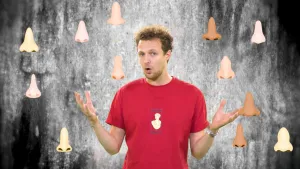 Een foto van Tim, die zijn handen met de handpalmen omhoog houdt. Vóór de foto zijn twaalf getekende neuzen te zien, die allemaal een andere vorm en kleur hebben. Zo heeft de ene neus brede neusvleugels, en de andere neus smalle neusvleugels.