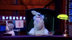 Een foto van pop Stella Schaap met een bord waarop een getekende vinger van een mens staat. De pop Nico de Neushoorn zit op een groene stoel met een bril op. Voor Nico op tafel liggen een notitieboek en pen en staan een kopje thee en een bureaulamp.