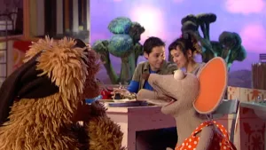 Een foto van de poppen Tommie en Ieniemienie die elkaar aankijken. Tommy met zwarte doek om zijn hoofd, Ieniemienie in rode blouse met witte stippen. Erachter zitten Elvan en Mamoun aan een houten tafel. Achteraan staan grote, groene stronken broccoli.