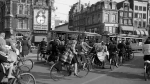 Een zwart-wit foto van mensen op de fiets in een Nederlandse straat in 1941. Rechtsachter rijdt een tram. Helemaal achteraan staan oude gebouwen. Op één staat "hotel", op een ander een klok met getekende man in pak erop en de tekst "Dobbelmann" eronder.