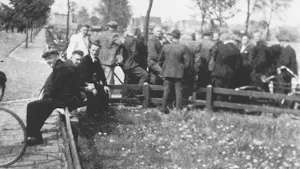Een zwart-wit foto van de April-meistaking in 1943 bij een laag hek om een grasveld. Rechts staan mannen achter het hek op een stoep en staat er een fiets tegenaan. Links zitten drie mannen op het hek en op de stoep is een voorwiel van een fiets te zien.