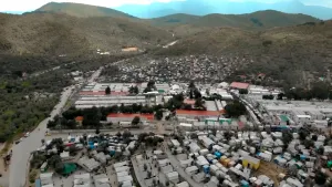 Een foto van het Griekse eiland Lesbos van bovenaf genomen. Er staan allemaal kleine huisjes, caravans en een enkele tent. De meeste zijn wit. Het zijn opvangplekken voor vluchtelingen. In de achtergrond zijn groenachtige heuvels of bergen te zien.