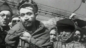 Een zwart-wit foto van mensen die achter prikkeldraad staan in een concentratiekamp van de nazi's. Ze kijken somber in de richting van de camera. Er zijn vier mannen en drie vrouwen te zien. Ze hebben allemaal een pet op of een doek om hun hoofd. 