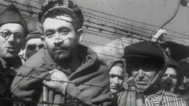 Een zwart-wit foto van mensen die achter prikkeldraad staan in een concentratiekamp van de nazi's. Ze kijken somber in de richting van de camera. Er zijn vier mannen en drie vrouwen te zien. Ze hebben allemaal een pet op of een doek om hun hoofd. 