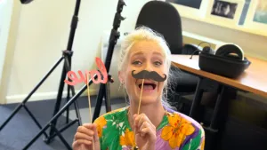 Een foto van presentator Janouk in een ruimte met bureau en bureaustoel. Ze heeft twee stokjes vast, één van een krullende zwarte snor die ze voor haar bovenlip houdt en één met het woord "love", liefde, in rode letters die in spiegelbeeld te zien zijn. 