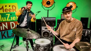 Een foto van Buddy met een microfoon in zijn hand in een groen met gele jas over een wit t-shirt. Hij is aan het rappen en gebaart met de vingers van zijn andere hand naar de camera. Ervoor zit meester Willem met houten drumstokken achter een drumstel.