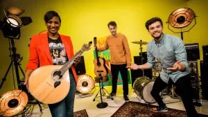Een foto van Edsilia Rombley met een portugese Gitaar met twaalf snaren. De kast van de gitaar heeft een bijna ronde vorm. Meester Willem heeft een gewone gitaar vast en Buddy staat met zijn handpalmen omhoog voor zijn lichaam. Ze kijken alledrie vrolijk.